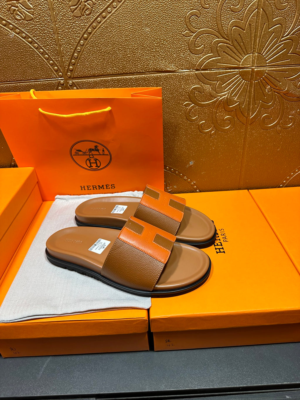 Tan slide sandals with orange H strap and black sole, resting on orange box.