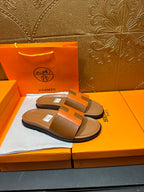 Tan slide sandals with orange H strap and black sole, resting on orange box.