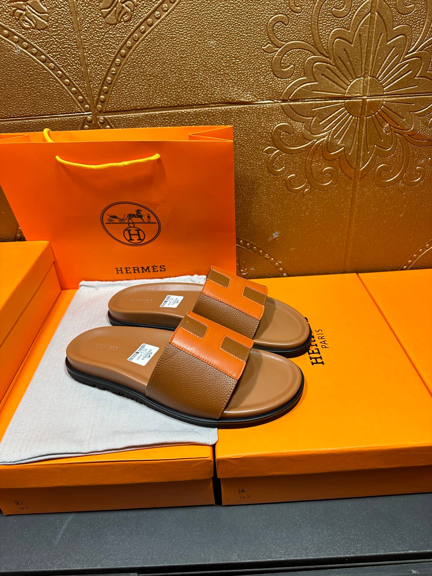 Tan slide sandals with orange H strap and black sole, resting on orange box.