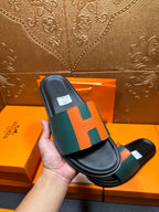 Hermes slide sandal held in hand, green strap with orange H blocks on black sole.