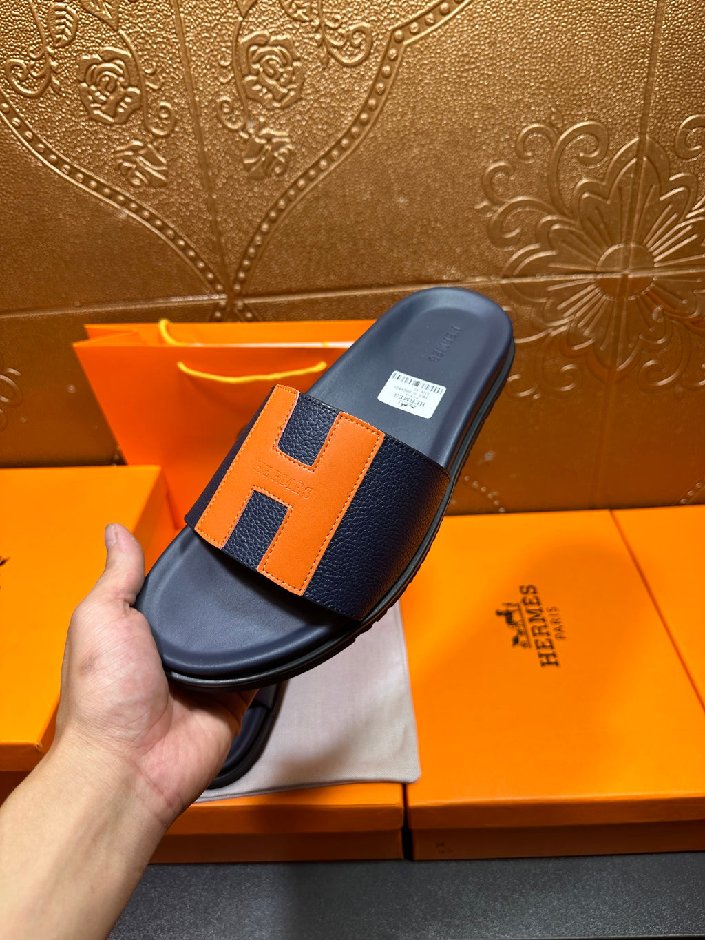Hermes slide sandal held by hand, dark navy with orange H straps and flat outsole