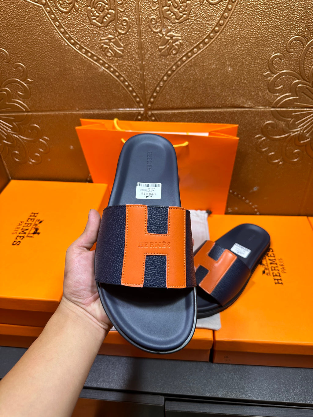 Hermes slide sandal held up, dark navy strap with orange H panel detail and footbed
