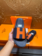 Hermes slide sandal held up, dark navy strap with orange H panel detail and footbed