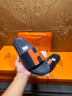 Hermes slide sandal held in hand, dark navy with orange H strap detail and flat sole