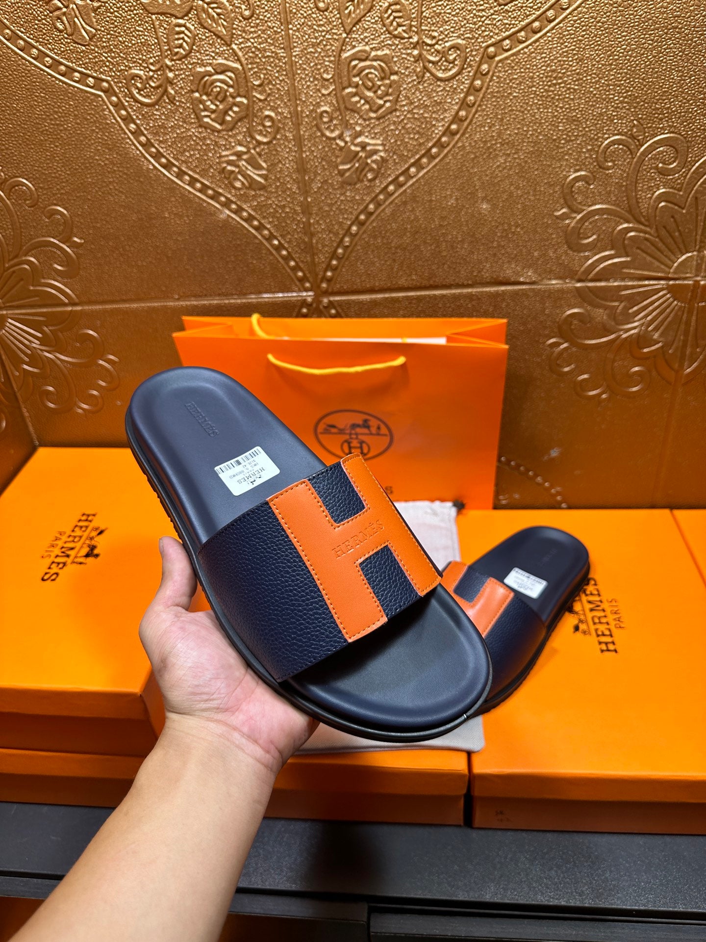 Hermes slide sandal held in hand, dark navy with orange H strap detail and flat sole