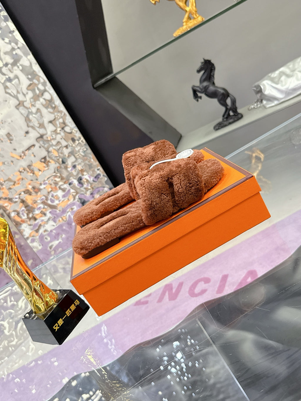 Brown plush slide sandals resting on an orange box, with white logo charm