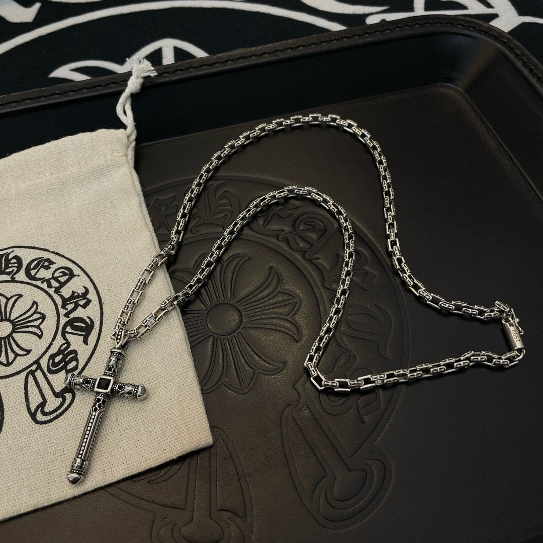Silver-tone cross pendant necklace draped on beige drawstring pouch over black box.