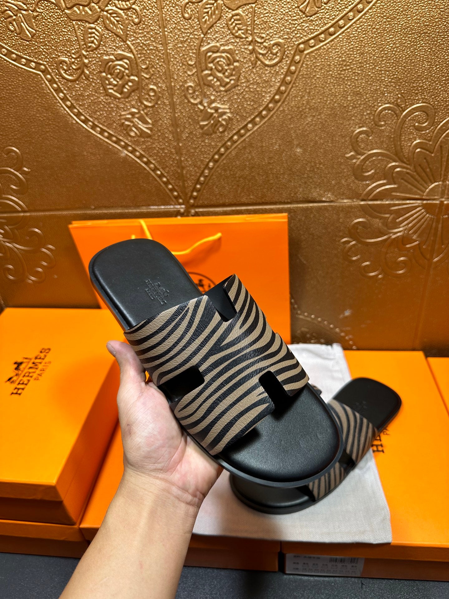 Hand holding black slide sandals with zebra-striped straps on white cloth.