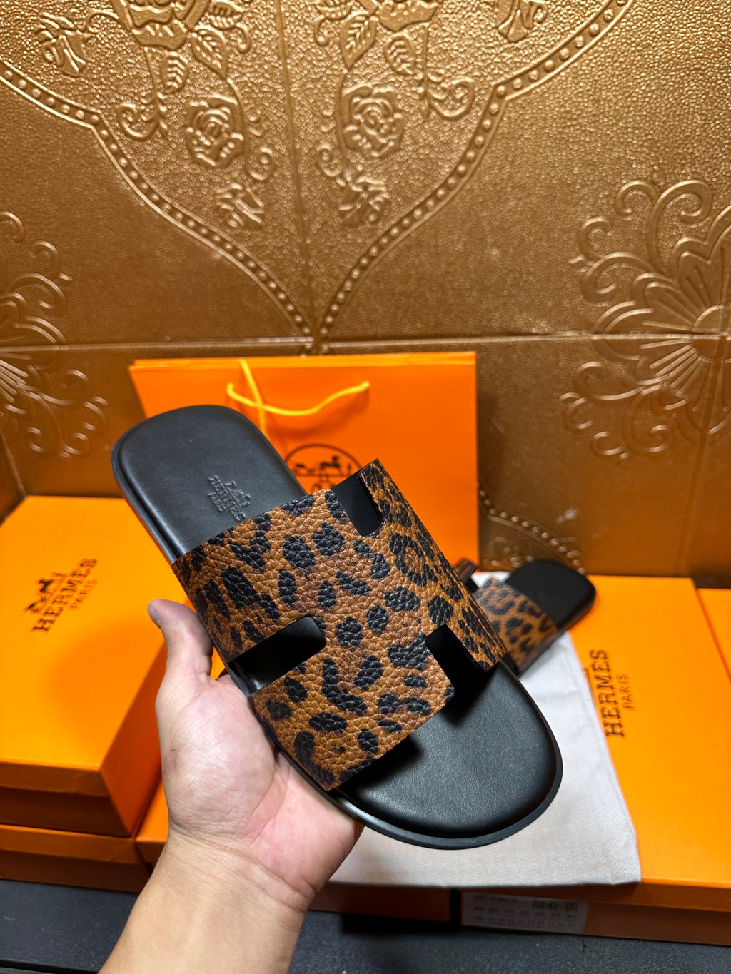 Hand holding leopard-print slide sandals with black rubber base and wide toe strap.