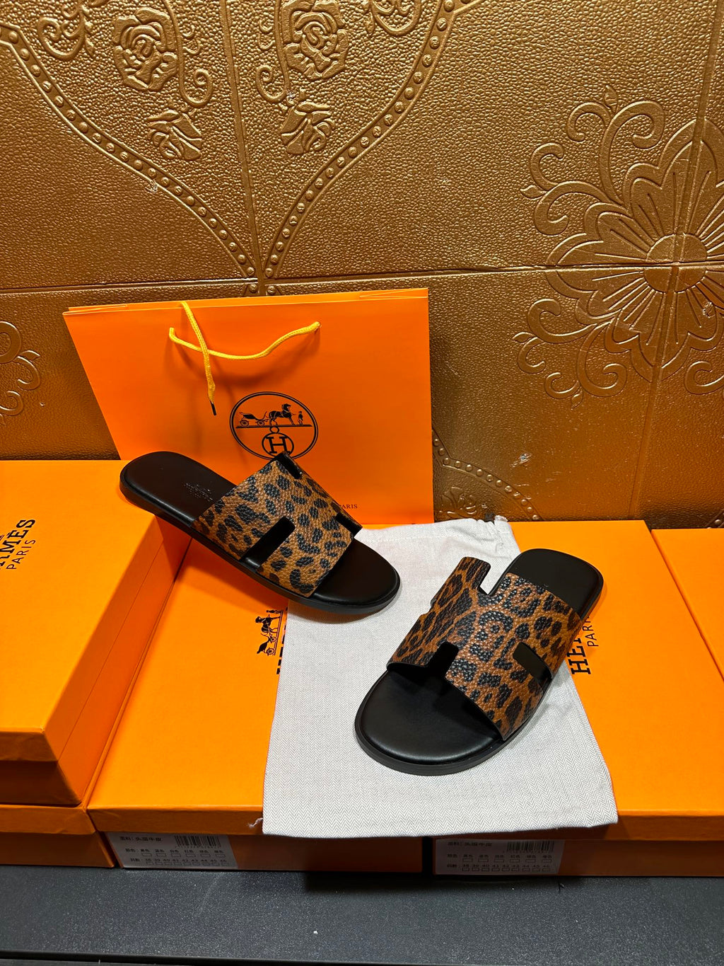 Leopard-print slide sandals with black soles, arranged on dust bag beside orange boxes.