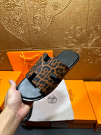 Leopard-print slide sandal on black footbed, held up over white dust bag and box.