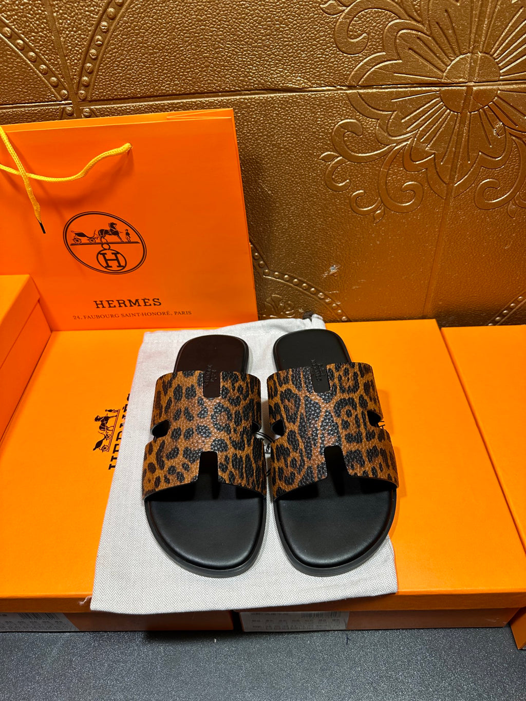 Leopard-print slide sandals with black straps and wide toe band, on white dust bag.