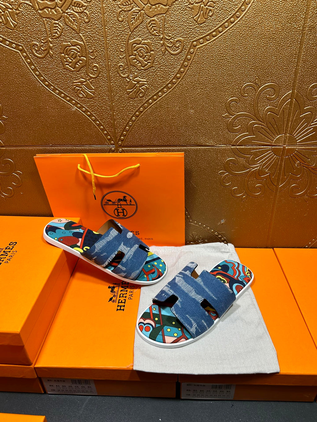Blue denim slide sandals with colorful artwork straps, held above orange box