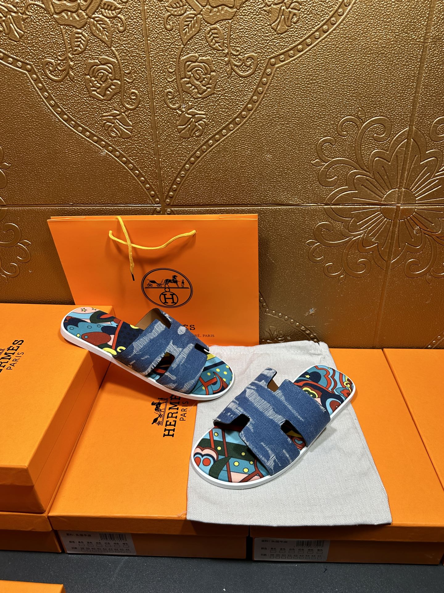 Blue denim slide sandals with colorful artwork straps, held above orange box