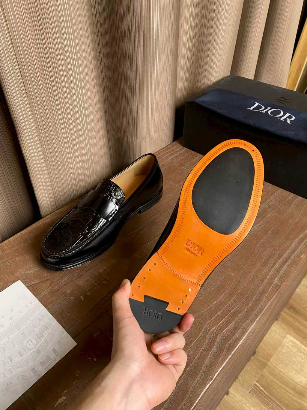 Black leather loafer beside orange insole with “DIOR” branding; hand holding
