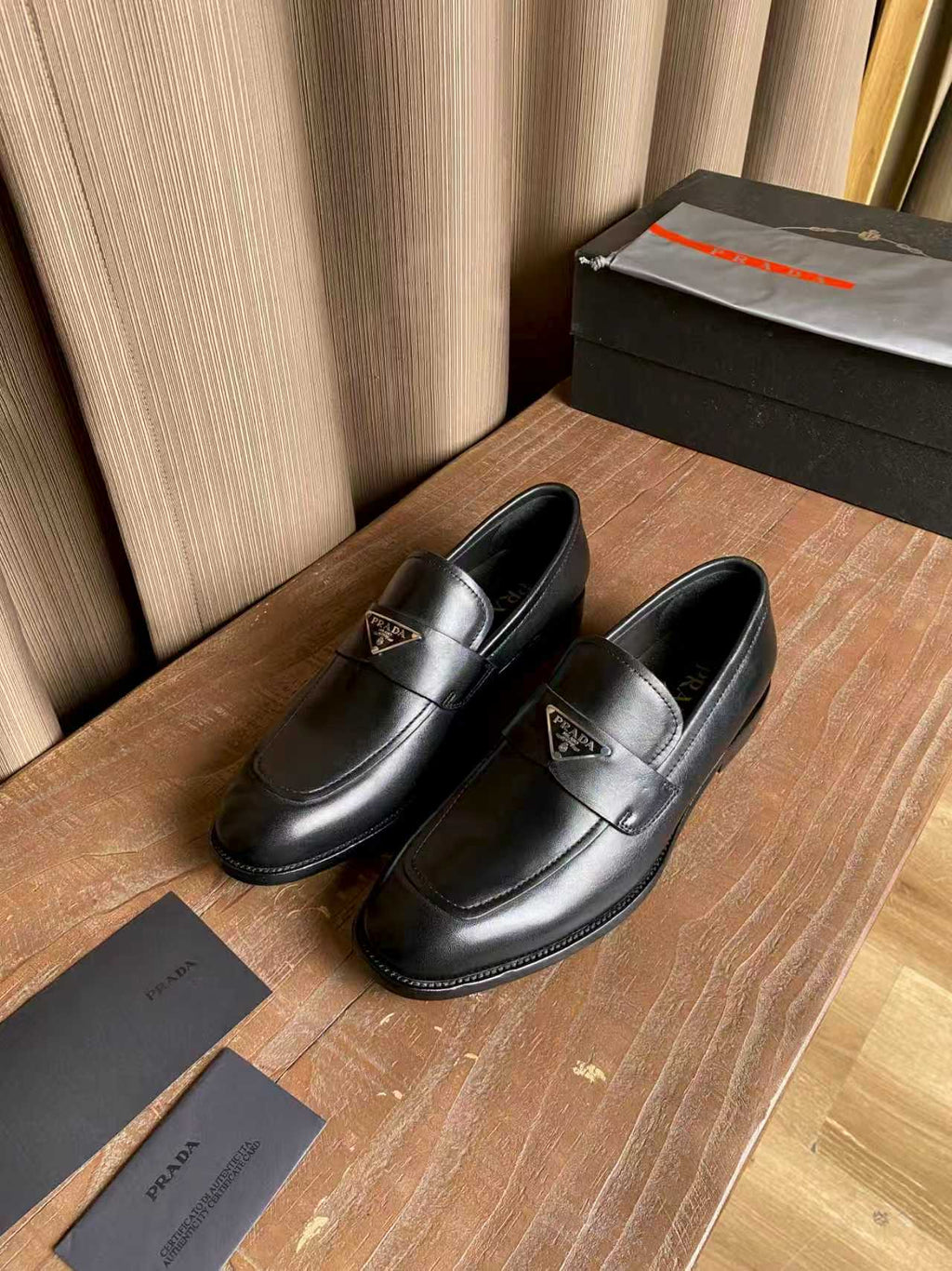 Prada black leather loafers, two pairs angled, strap and stitched apron visible