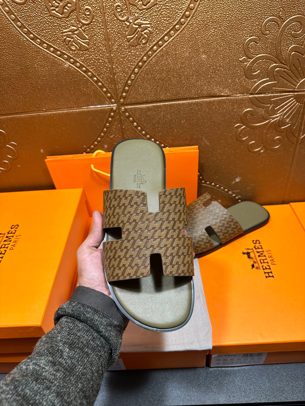 Hand holding Hermes slide sandals with dark patterned straps and light gray footbeds