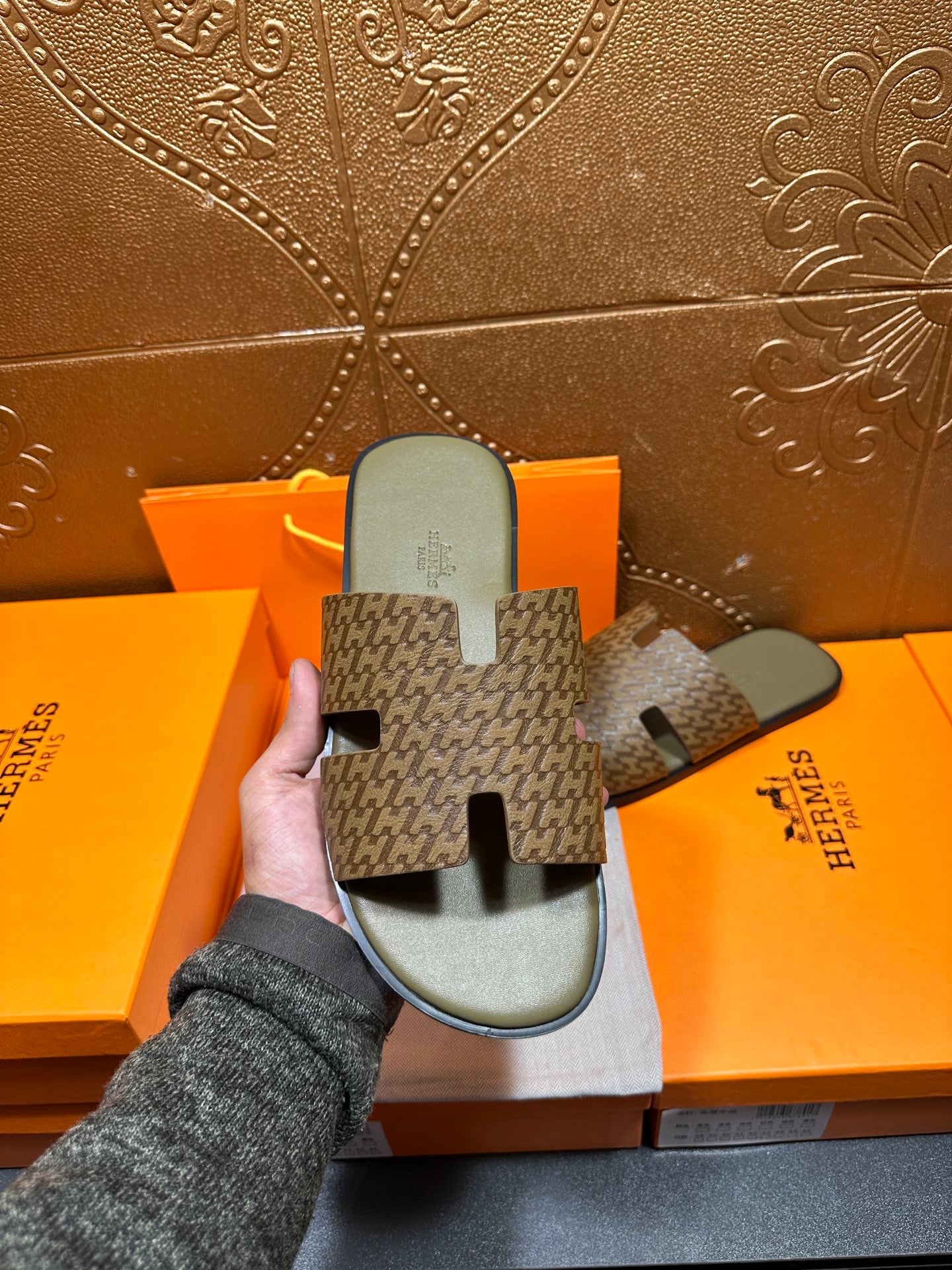 Hand holding Hermes slide sandals with dark patterned straps and light gray footbeds