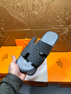 Hermes slide sandal held above box, dark patterned upper with black rubber footbed