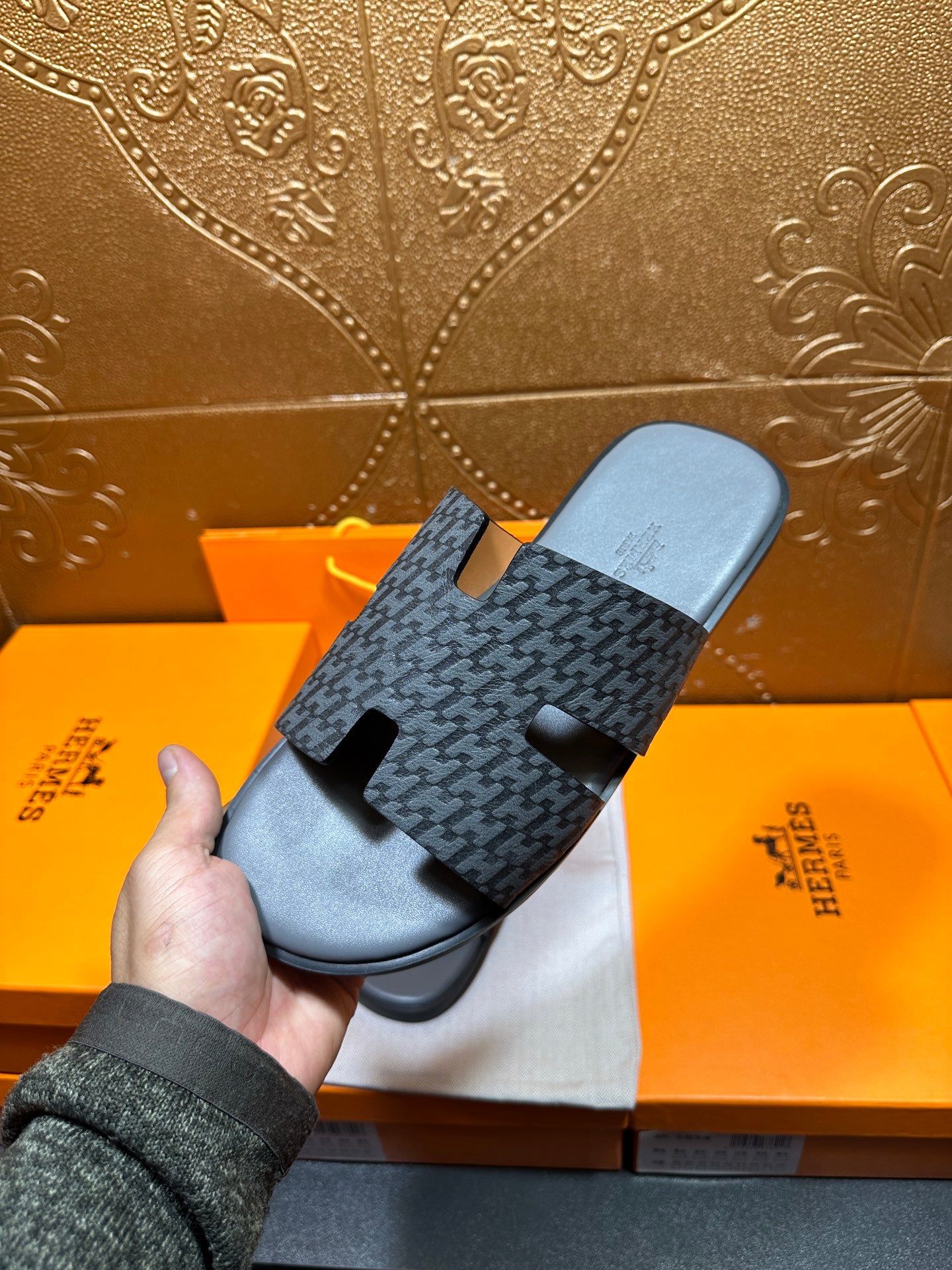 Hermes slide sandal held above box, dark patterned upper with black rubber footbed