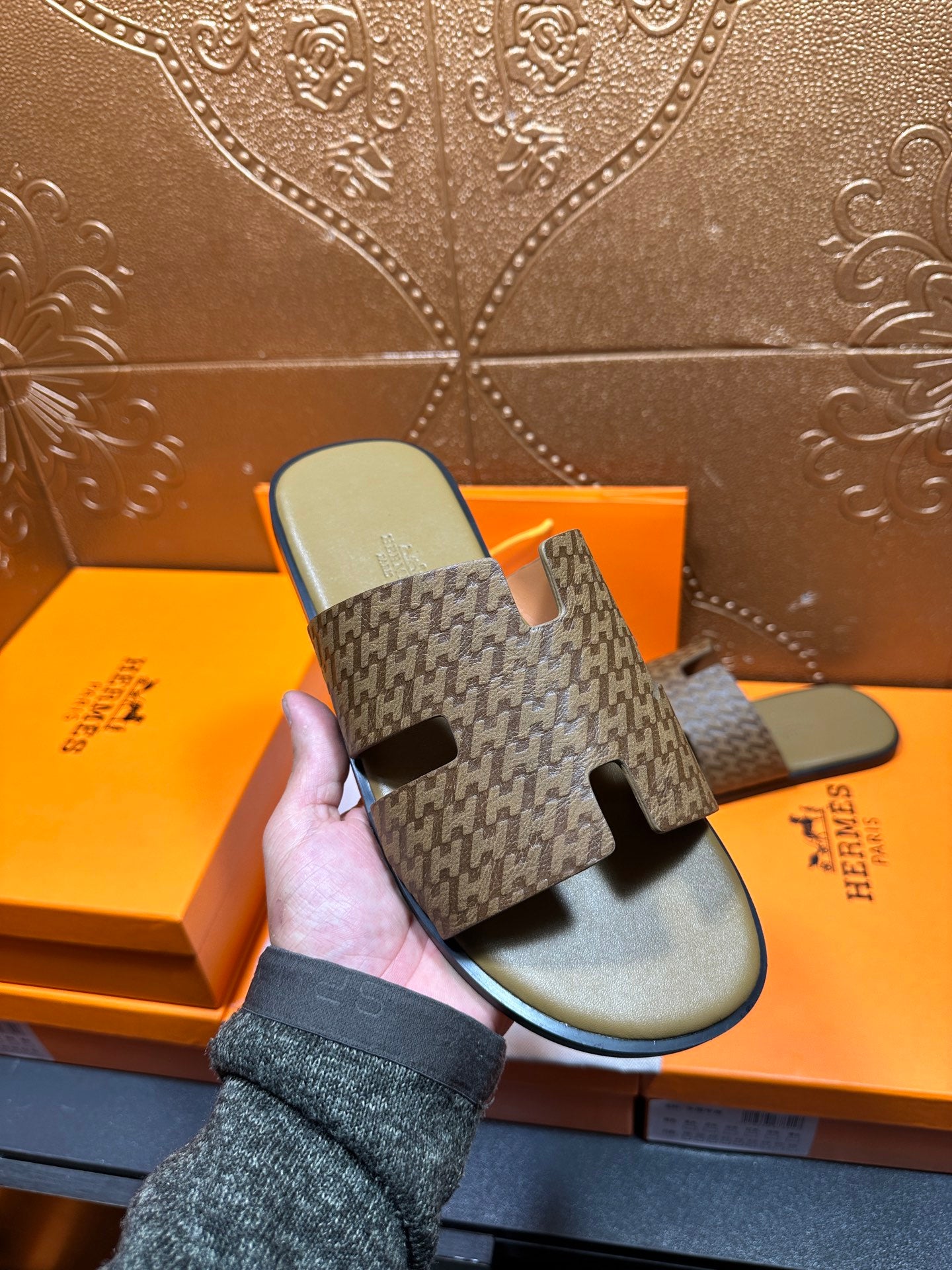 Hand holding Hermes slide sandals; patterned upper with black cutout details on olive footbeds