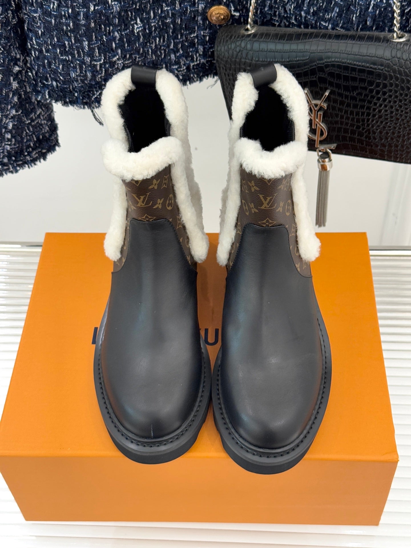 Two black ankle boots featuring white shearling trim and brown monogram side panels