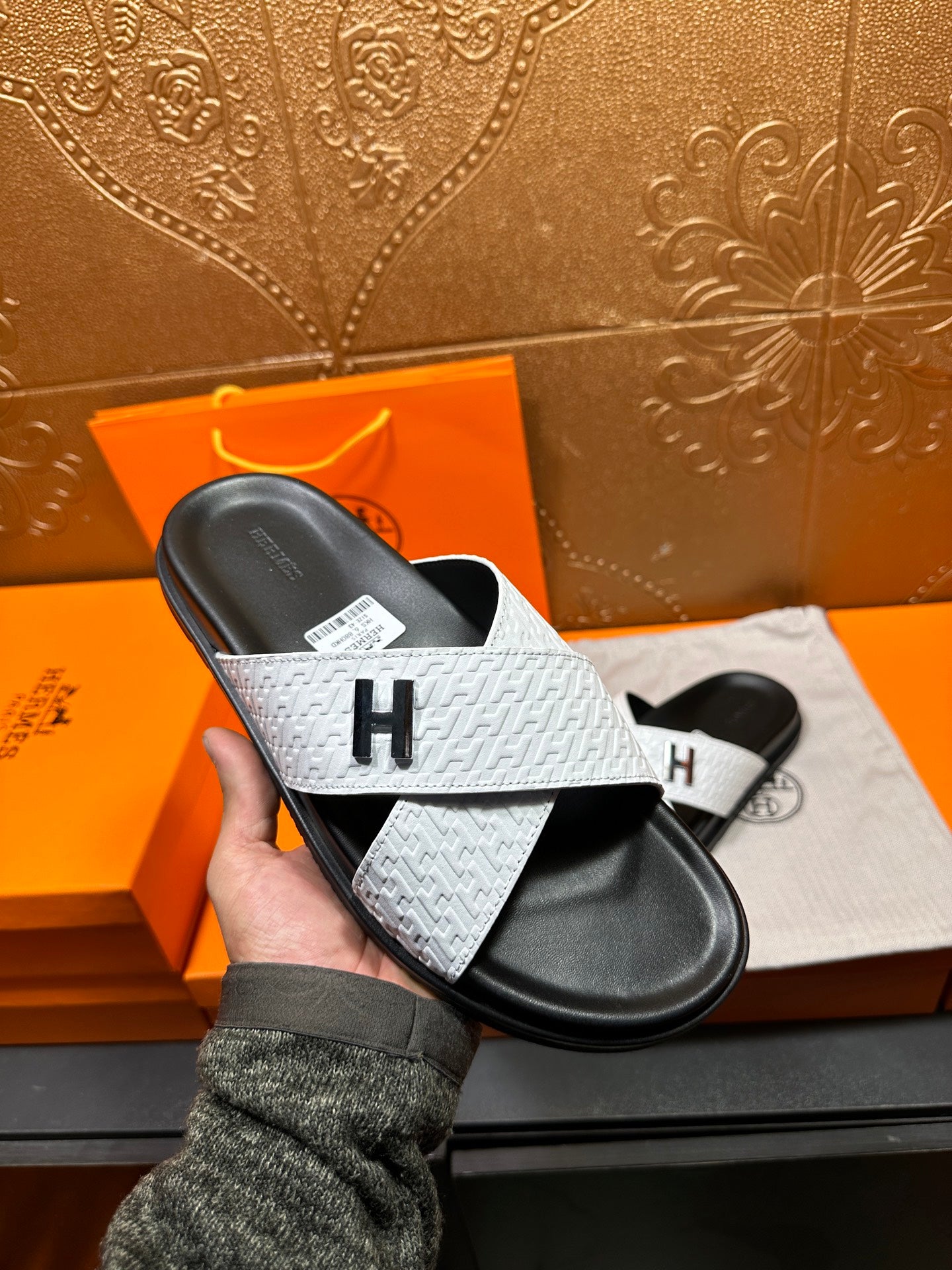 Hand holding a white textured slide sandal with black footbed and H on strap