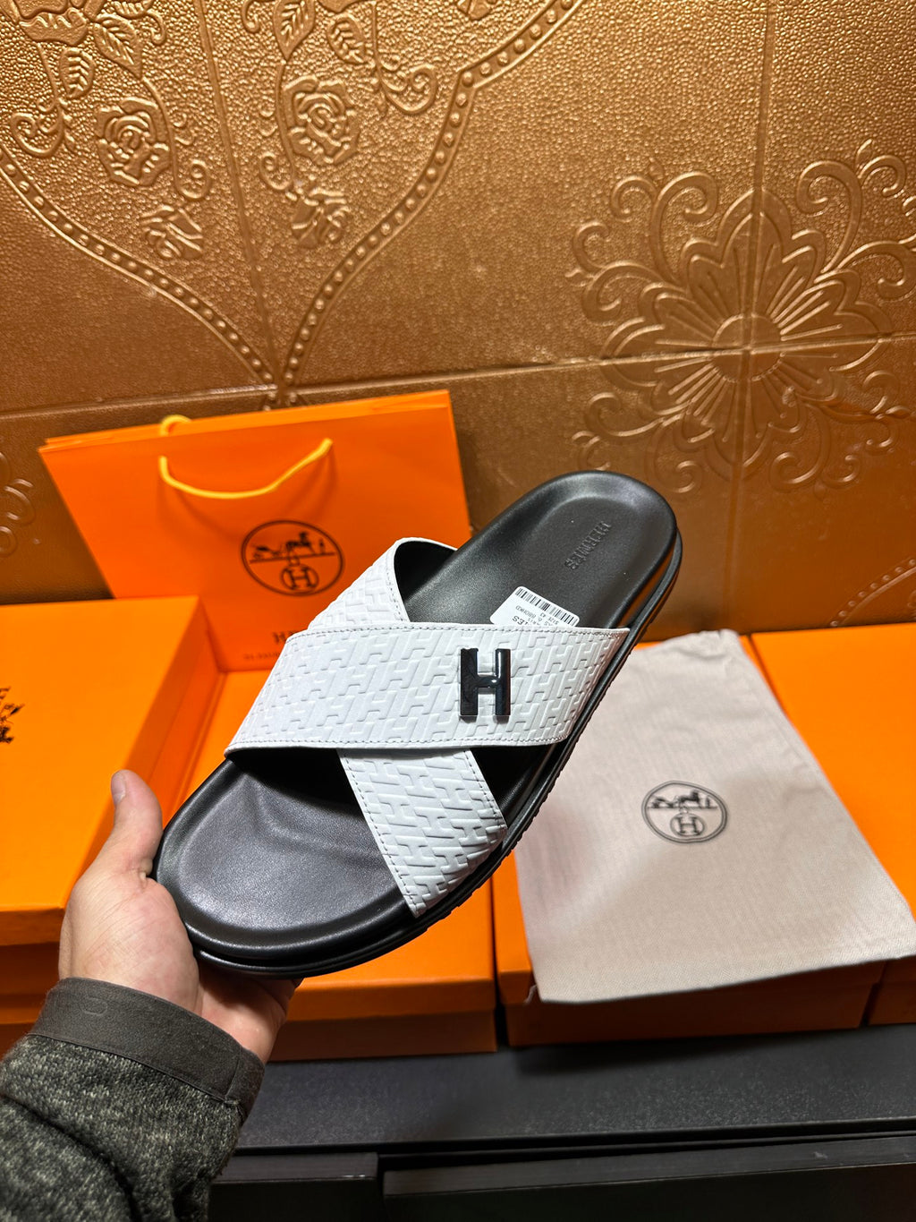 White textured slide sandal with black footbed and H emblem, held above dust bag