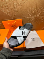 White textured slide sandal with black footbed and H emblem, held above dust bag