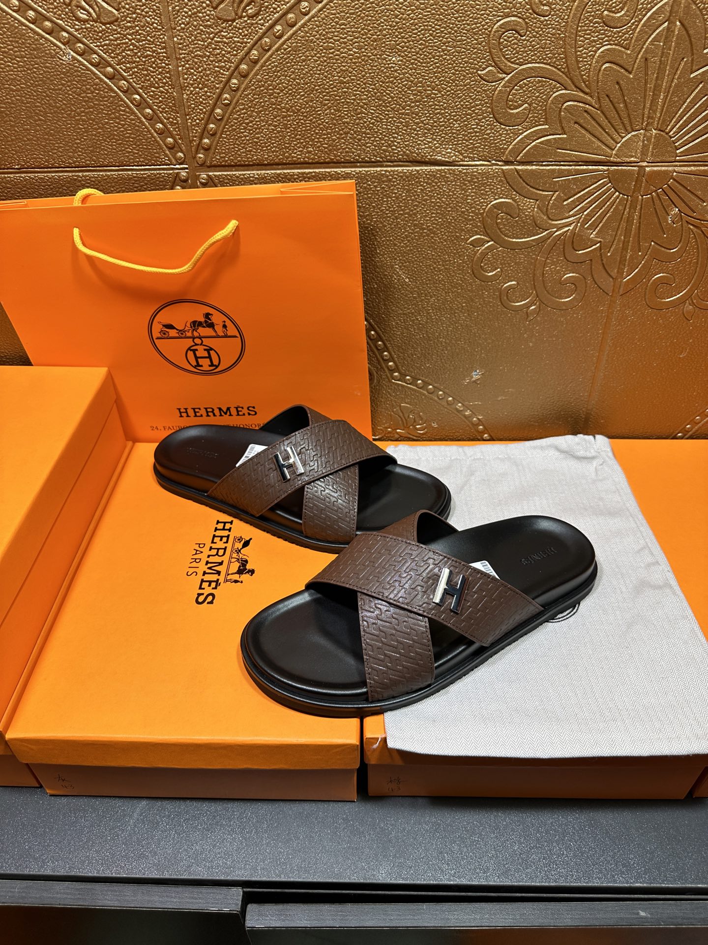 Pair of brown slide sandals with black footbed and H logo strap on a tray