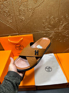 Hermes slide sandal held up, tan textured cross strap with H buckle, open toe