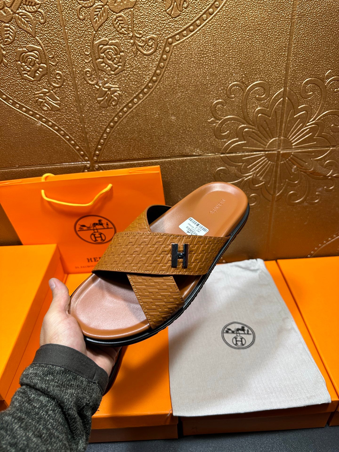 Hermes slide sandal held up, tan textured cross strap with H buckle, open toe