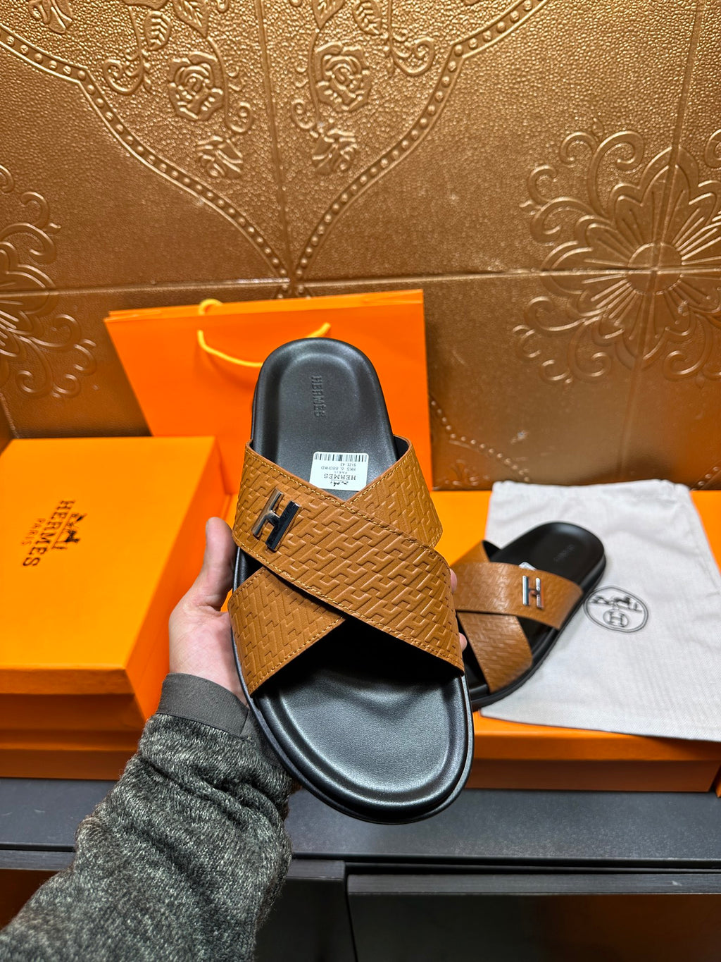 Brown textured leather slide sandals held in hand, black footbed and H logo