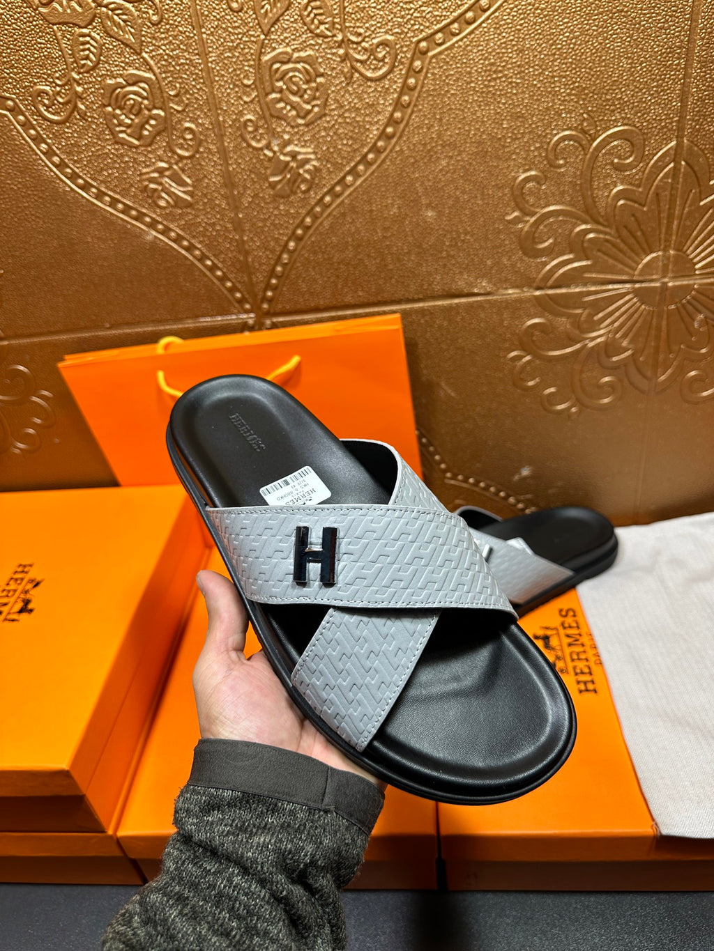 Hermes sandals being held: light gray strap over black footbed with H buckle