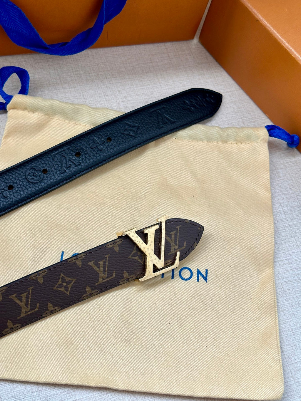 Monogram belt with dark strap and gold-tone LV buckle on a beige pouch