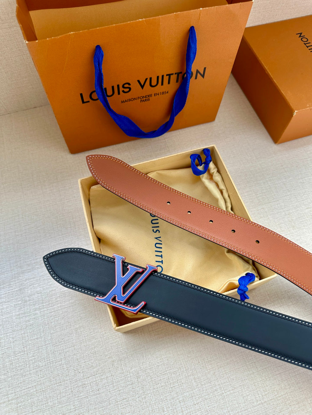 Two leather belts in box: dark blue and tan, each with LV monogram buckle