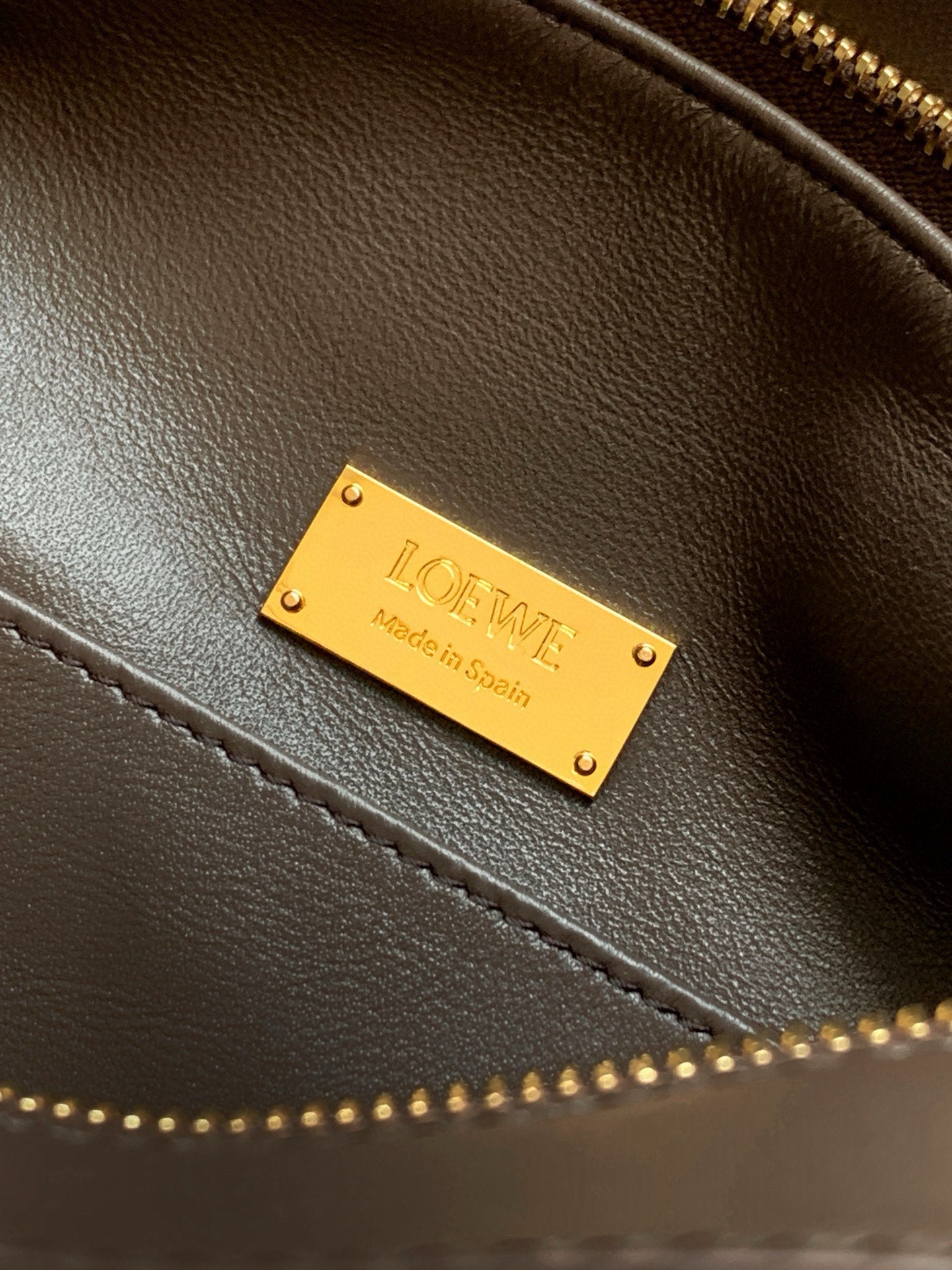 Leather bag close-up of gold logo plate and stitched trim, dark brown