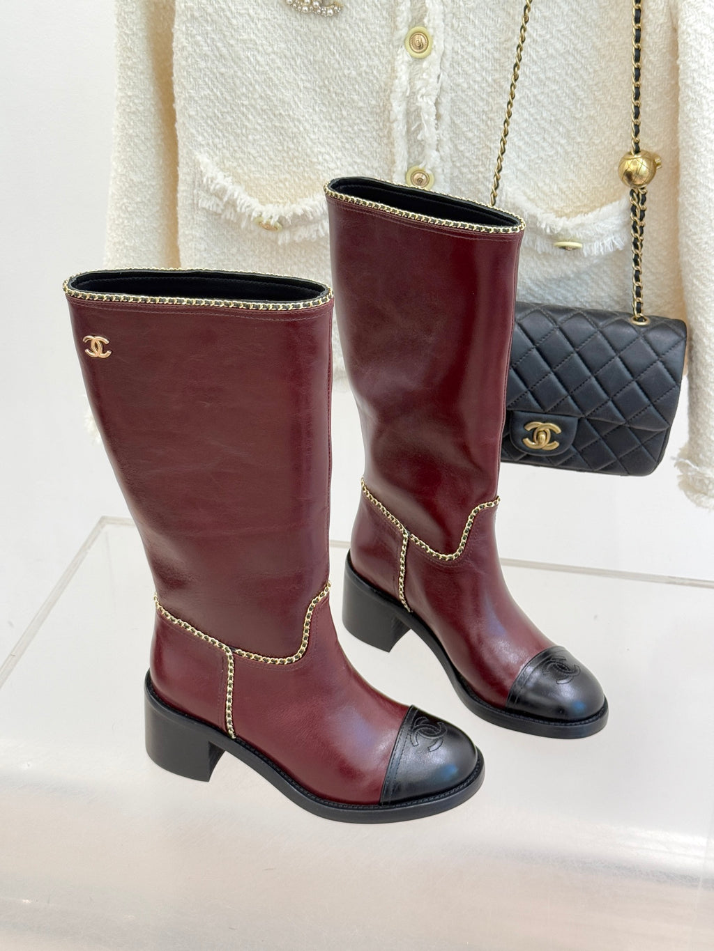 Two burgundy knee-high boots with black toe caps and gold chain trim