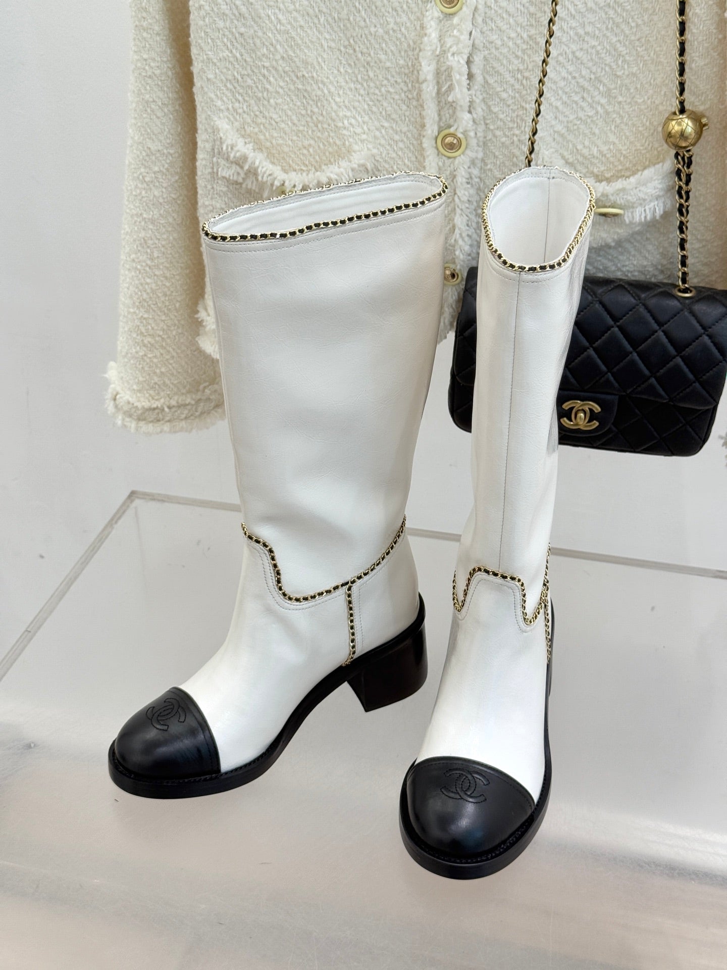 White leather knee boots with black toe caps, mid heel, and gold chain trim