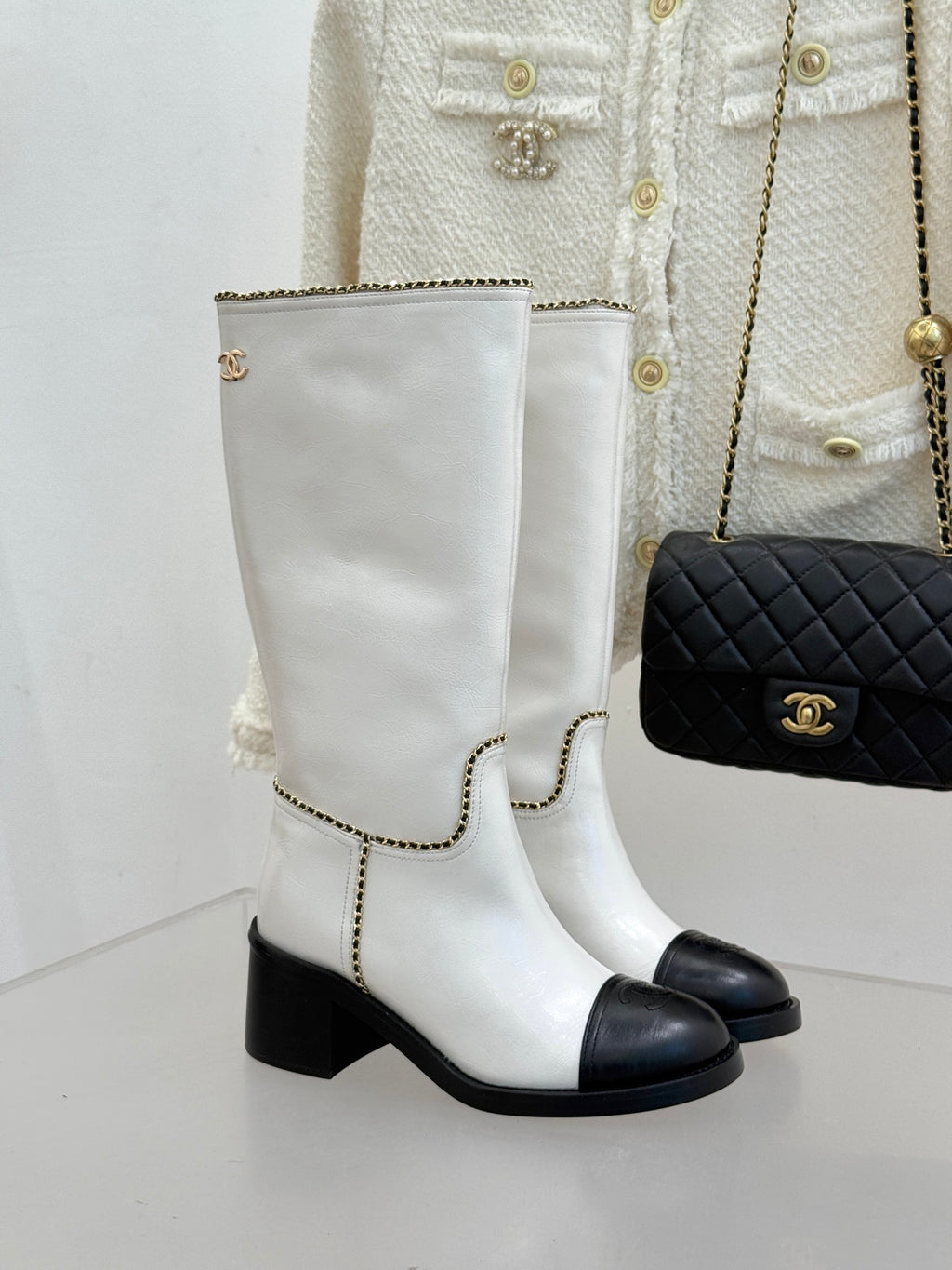 White leather knee boots with black toe panels, chunky heel, and gold chain trim