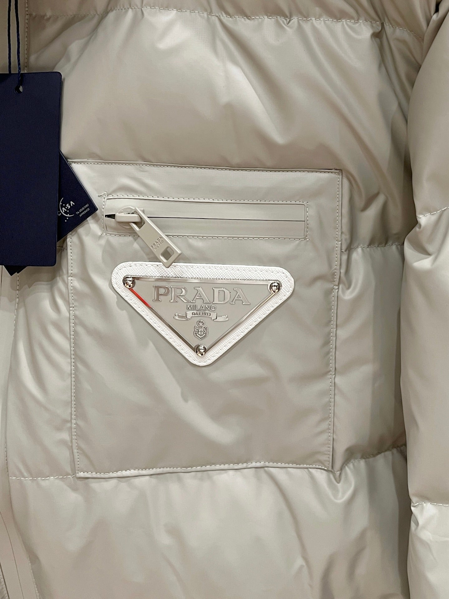 Long puffer jacket panel in light beige with rectangular pocket and triangular logo metal plaque