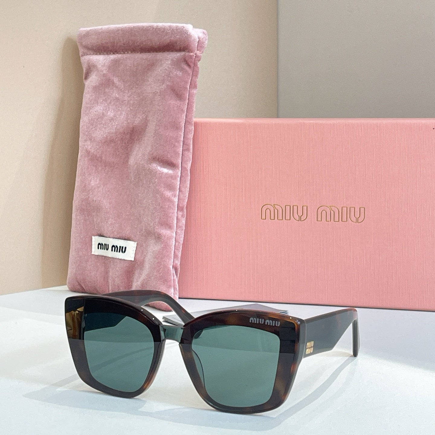 MiuMiu square sunglasses with dark frames and green-tinted lenses, in pink pouch