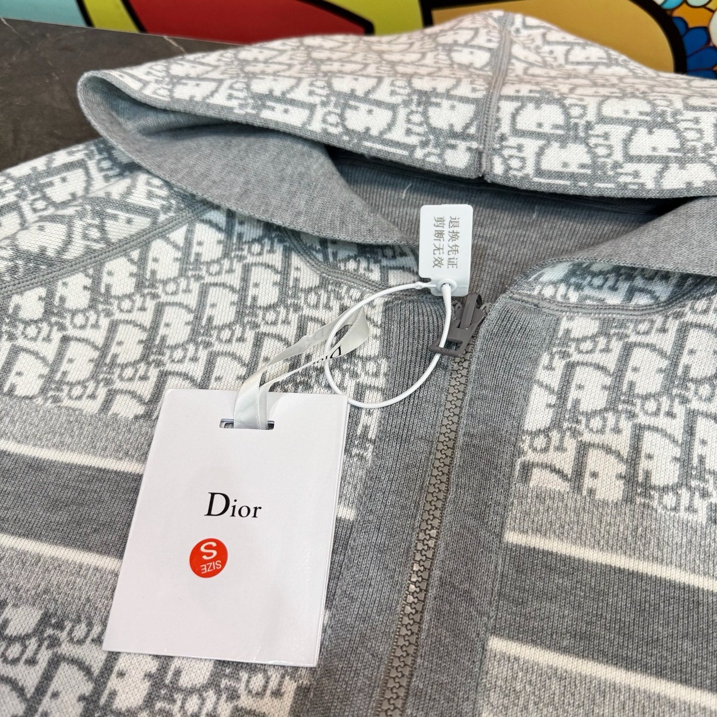 Close-up: gray and white monogram hooded sweatshirt fabric with Dior tag on zipper.