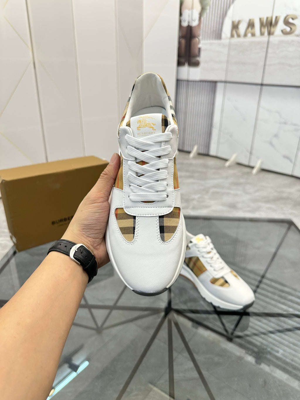 Burberry white lace-up sneaker held in hand, gold-toned trim on side panel