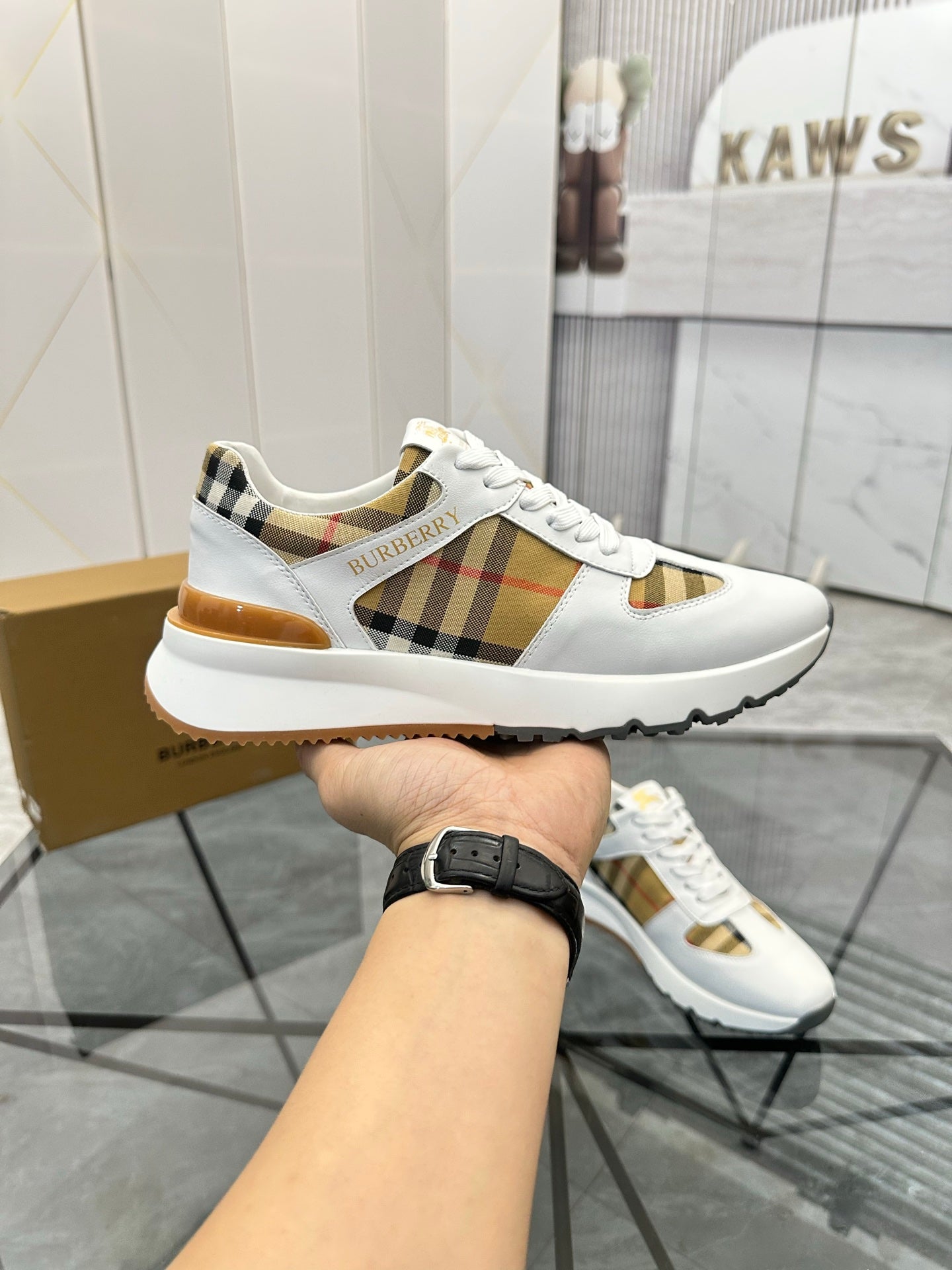 Burberry white sneaker held by a person, plaid side panel and gold-toned trim visible