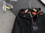 Long black winter jacket close-up: hood, zip front, orange piping, logo patch