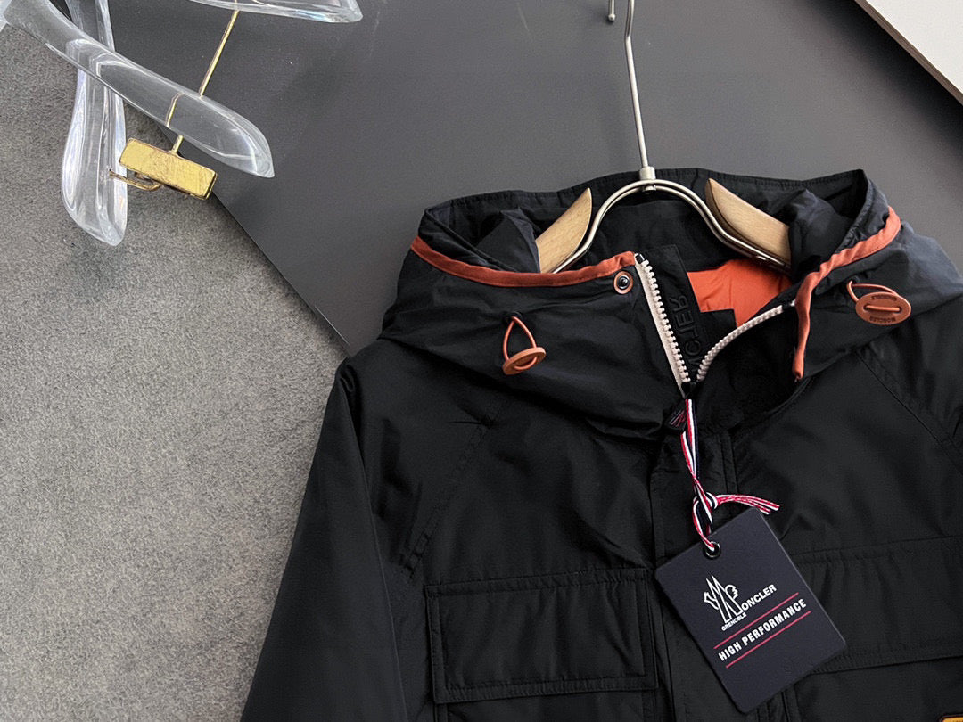 Long black winter jacket close-up: hood, zip front, orange piping, logo patch
