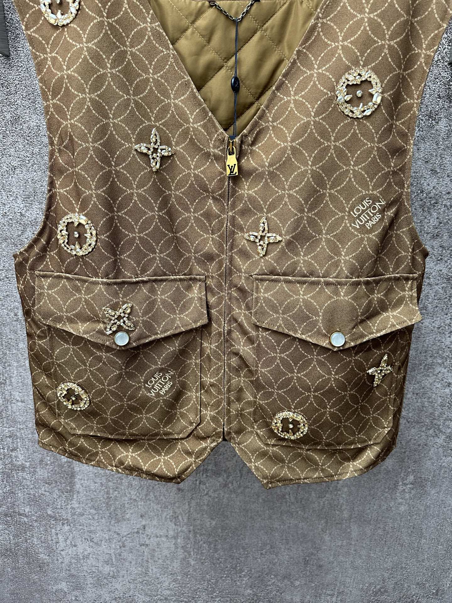 Gold monogram sleeveless vest with zip front, flap pockets, and small decorative charms