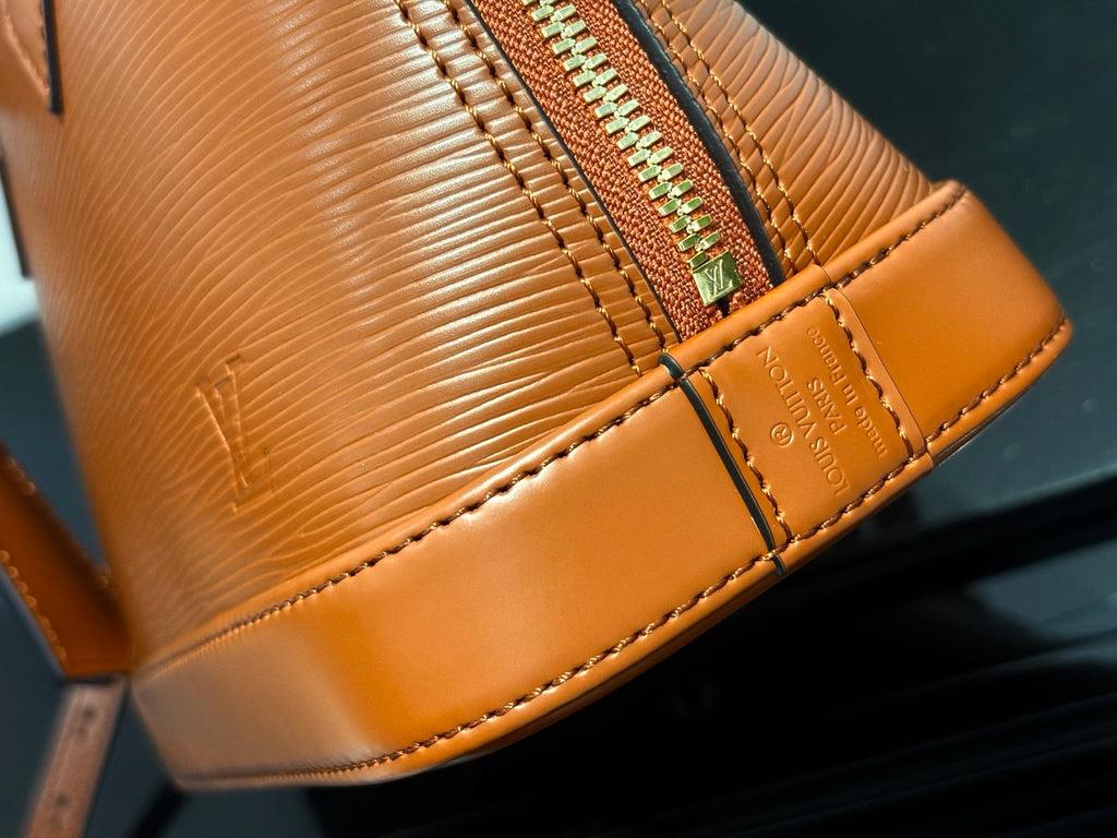 Tan leather handbag bottom corner with gold-trim stitching and embossed logo panel