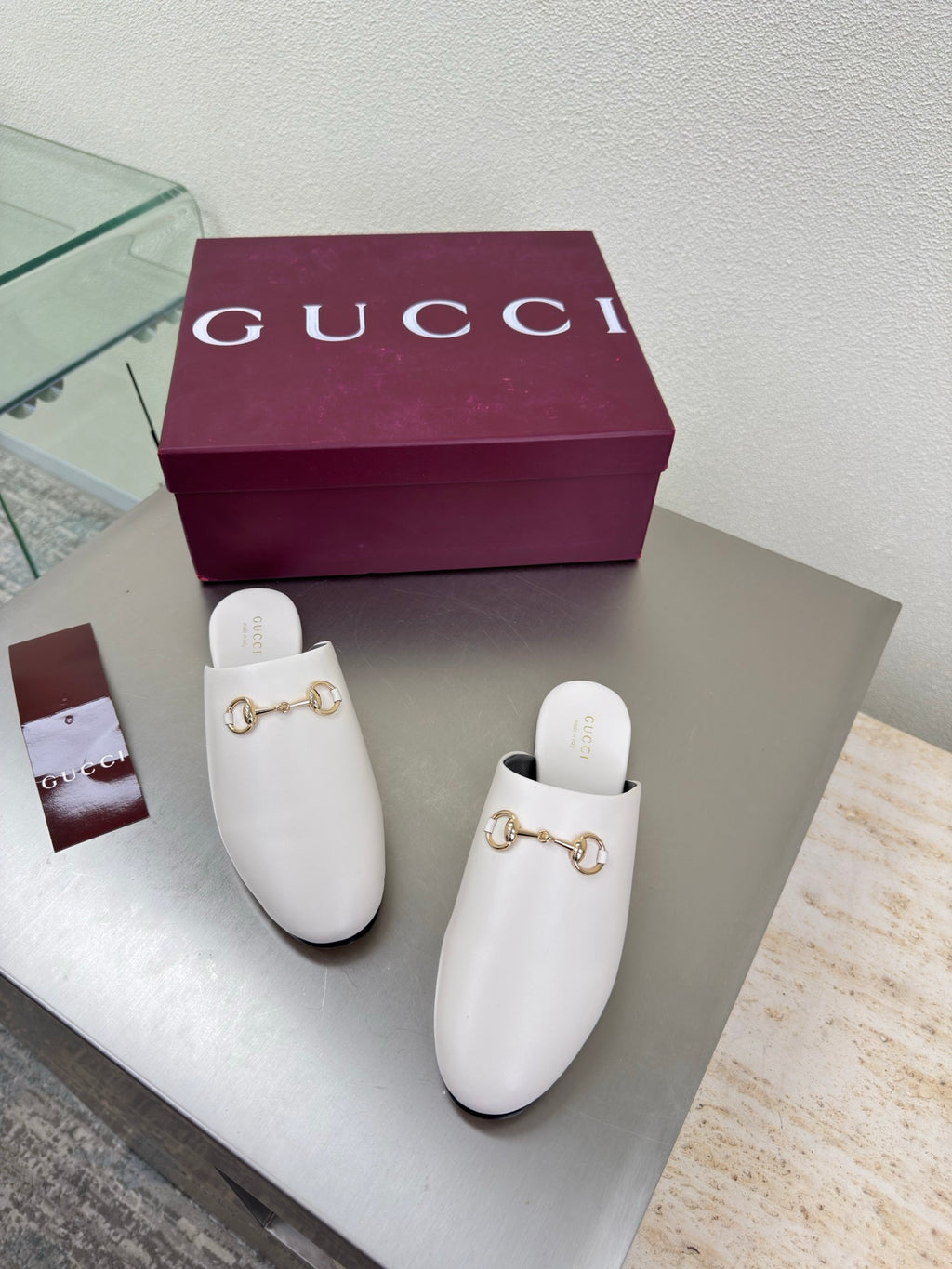 White mule shoes with gold horsebit hardware, pair displayed beside Gucci box